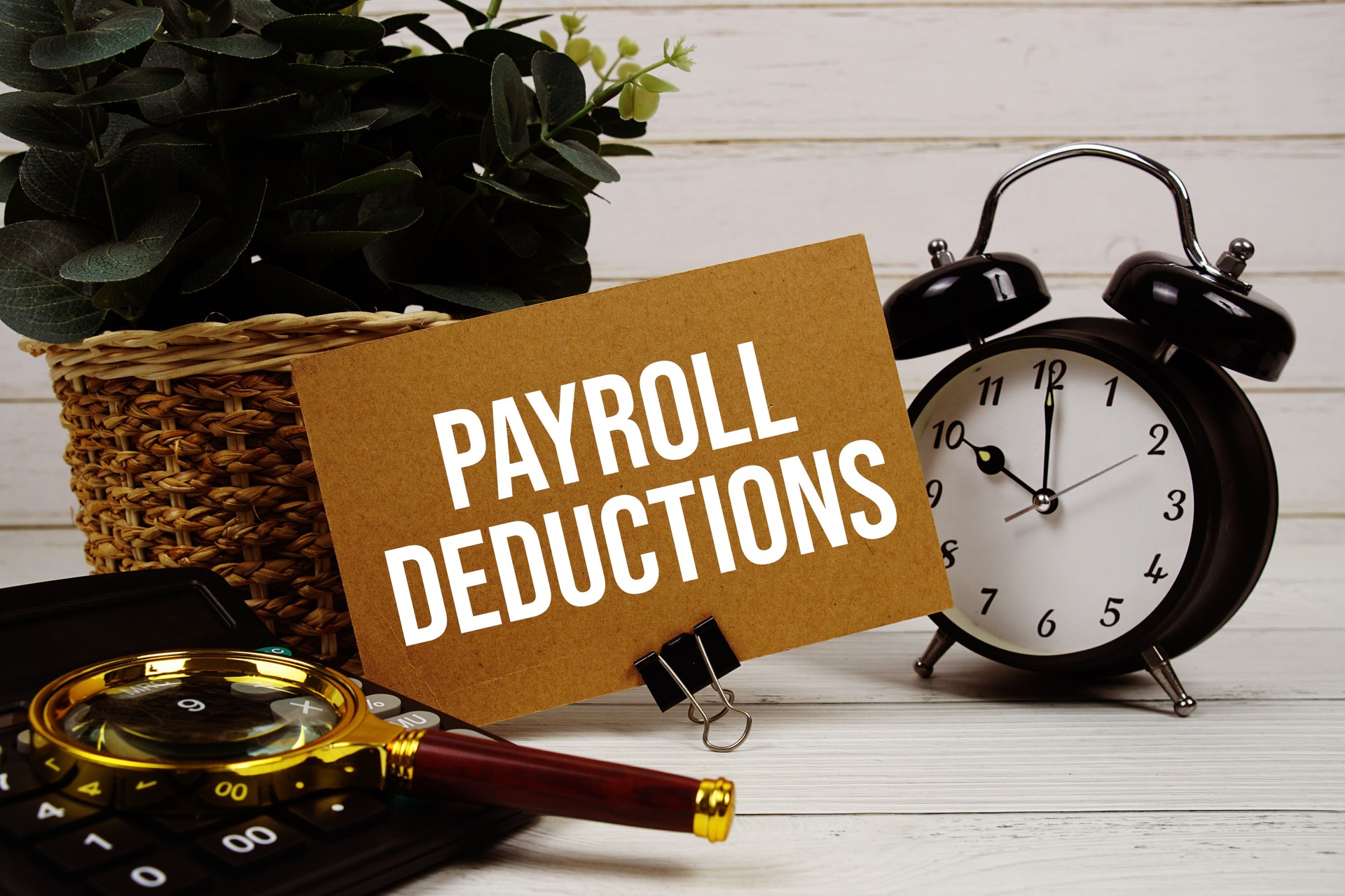 Payroll Deductions text on paper card with alarm clock, calculator and magnifying glass on wooden background